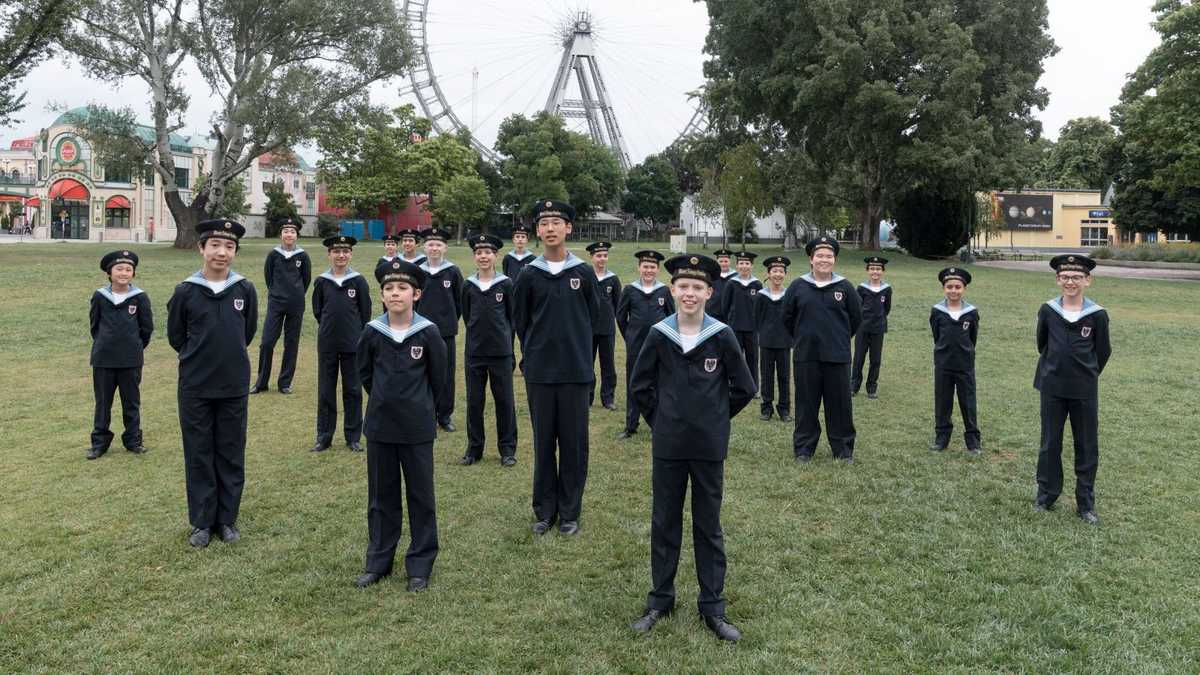 The Vienna Boys Choir standing outdoors at a park with a Ferris Wheel in the background
